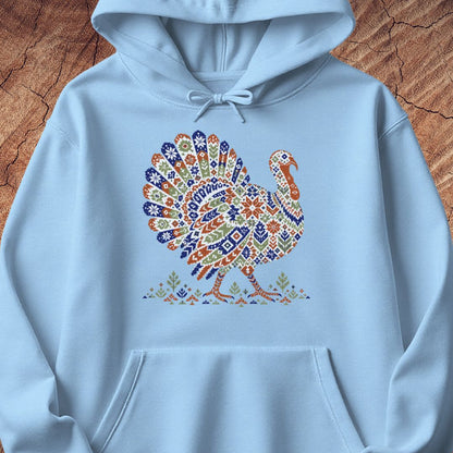 Light blue hoodie with a wild turkey patterned like a colorful knit sweater with snowflakes, stars, and tiny pine trees, laid flat on a rustic wood background.