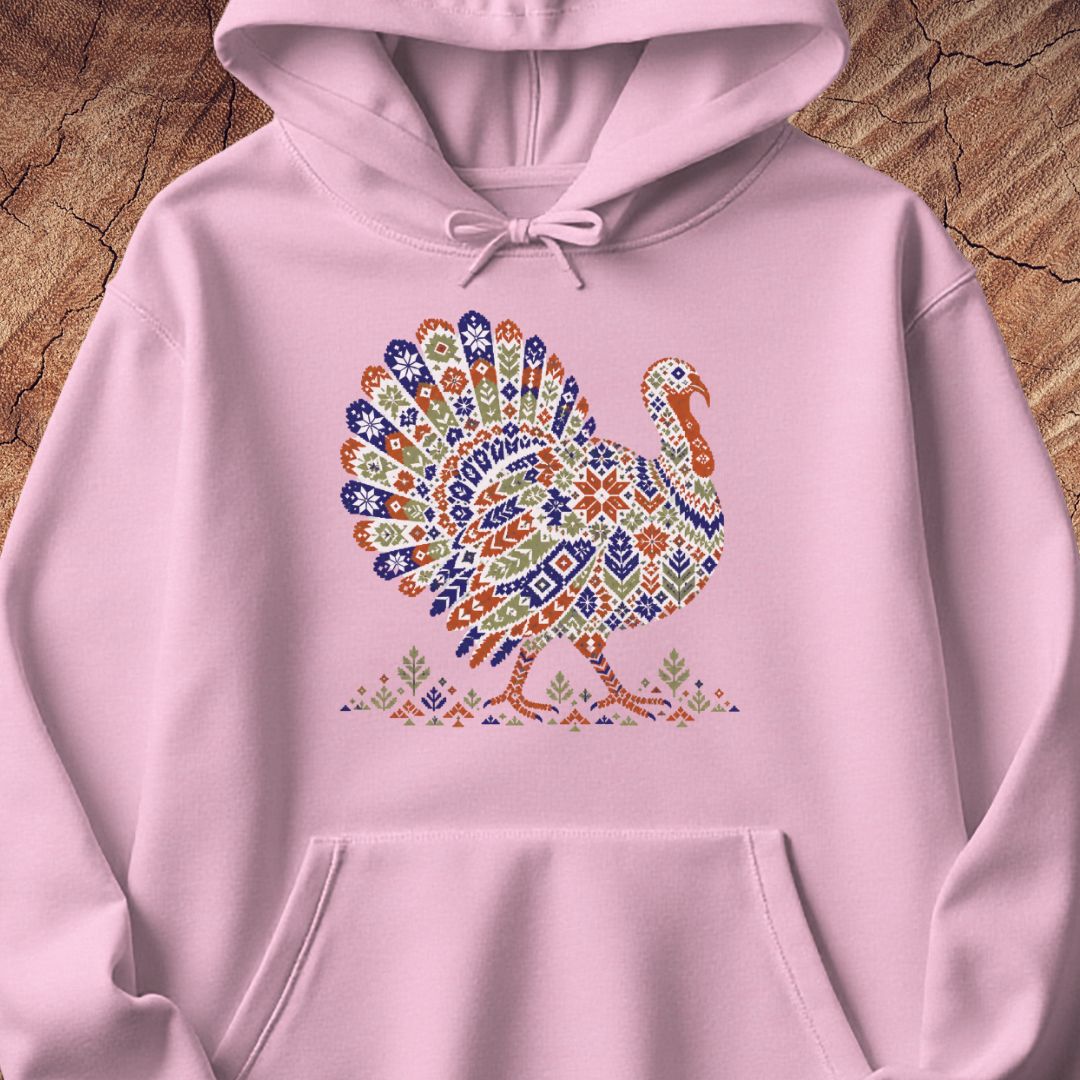 Light pink hoodie with a wild turkey patterned like a colorful knit sweater with snowflakes, stars, and tiny pine trees, laid flat on a rustic wood background.