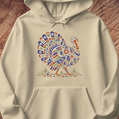 Sand beige hoodie with a wild turkey patterned like a colorful knit sweater with snowflakes, stars, and tiny pine trees, laid flat on a rustic wood background.