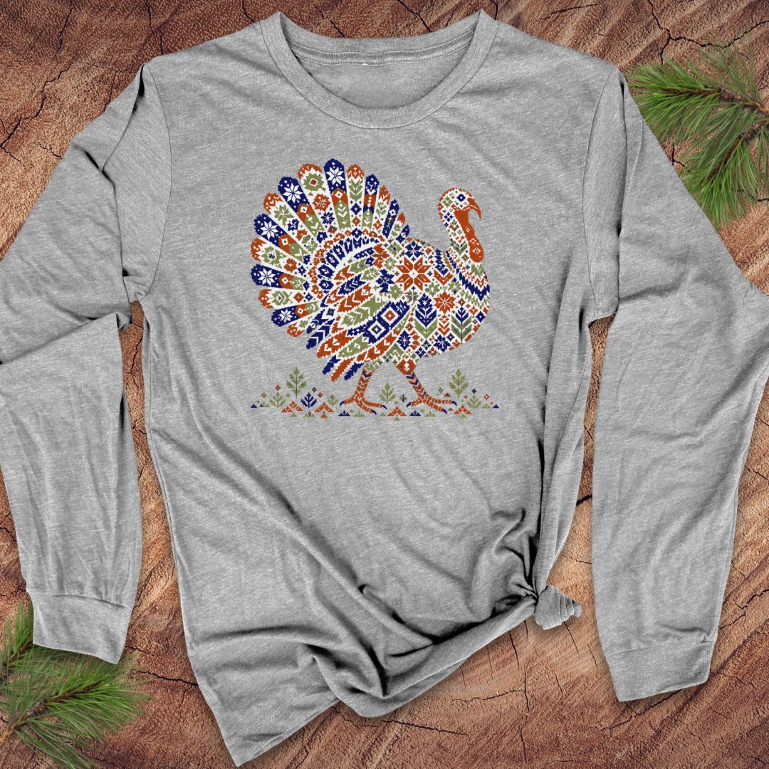 Athletic heather gray long-sleeve t-shirt with a wild turkey patterned like a colorful knit sweater with snowflakes, stars, and tiny pine trees, laid flat on a rustic wood background with pine branches.
