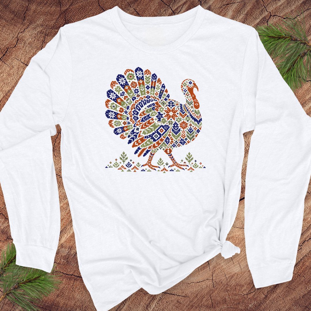 White long-sleeve t-shirt with a wild turkey patterned like a colorful knit sweater with snowflakes, stars, and tiny pine trees, laid flat on a rustic wood background with pine branches.