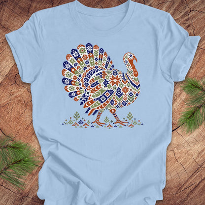 A light blue short-sleeve crewneck t-shirt featuring a colorful, patterned graphic of a Thanksgiving turkey in the center, laid flat on a rustic wood background with pine branches in the corners.