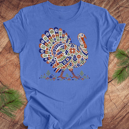 A blue short-sleeve crewneck t-shirt featuring a colorful, patterned graphic of a turkey on the chest, laid flat on a rustic wood background with pine branches.