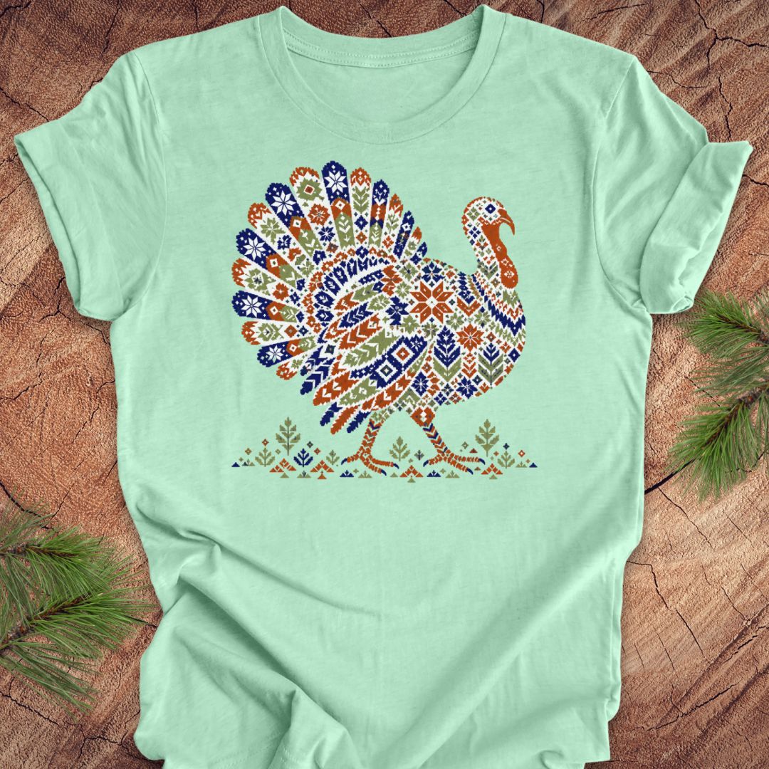 A mint green short-sleeve t-shirt with a colorful, patterned graphic of a turkey on the chest, laid flat on a rustic wood background with pine branches in the corners.