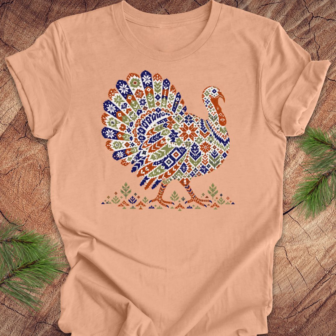 A peach-colored short-sleeve t-shirt with a colorful, patterned graphic of a Thanksgiving turkey, laid flat on a rustic wood background with pine branches.