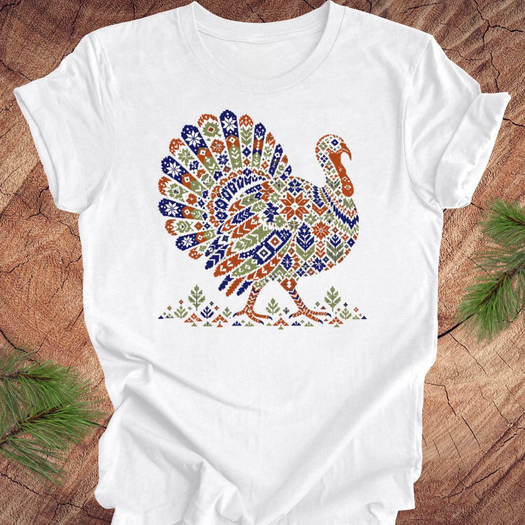 A white short-sleeve crewneck t-shirt featuring a colorful, patterned graphic of a turkey on the chest. The shirt is laid flat on a rustic wood background with pine branches visible in the corners.