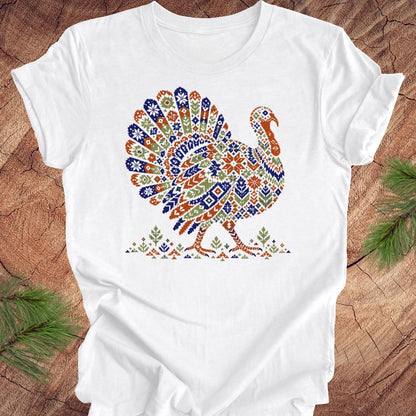A white short-sleeve crewneck t-shirt featuring a colorful, patterned graphic of a turkey on the chest. The shirt is laid flat on a rustic wood background with pine branches visible in the corners.