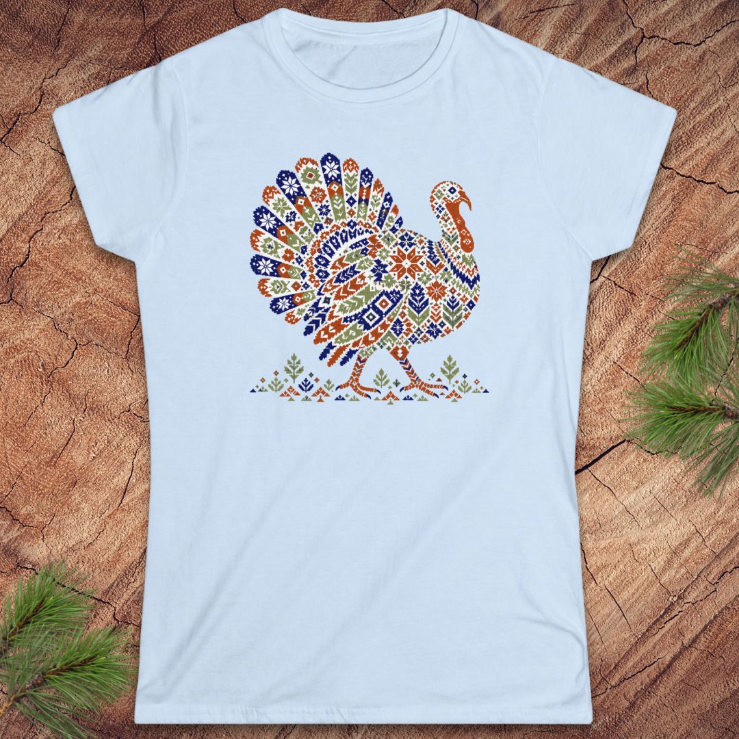 Light blue women's t-shirt with a wild turkey patterned like a colorful knit sweater with snowflakes, stars, and tiny pine trees, laid flat on a rustic wood background with pine branches.
