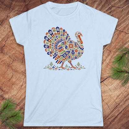 Light blue women's t-shirt with a wild turkey patterned like a colorful knit sweater with snowflakes, stars, and tiny pine trees, laid flat on a rustic wood background with pine branches.