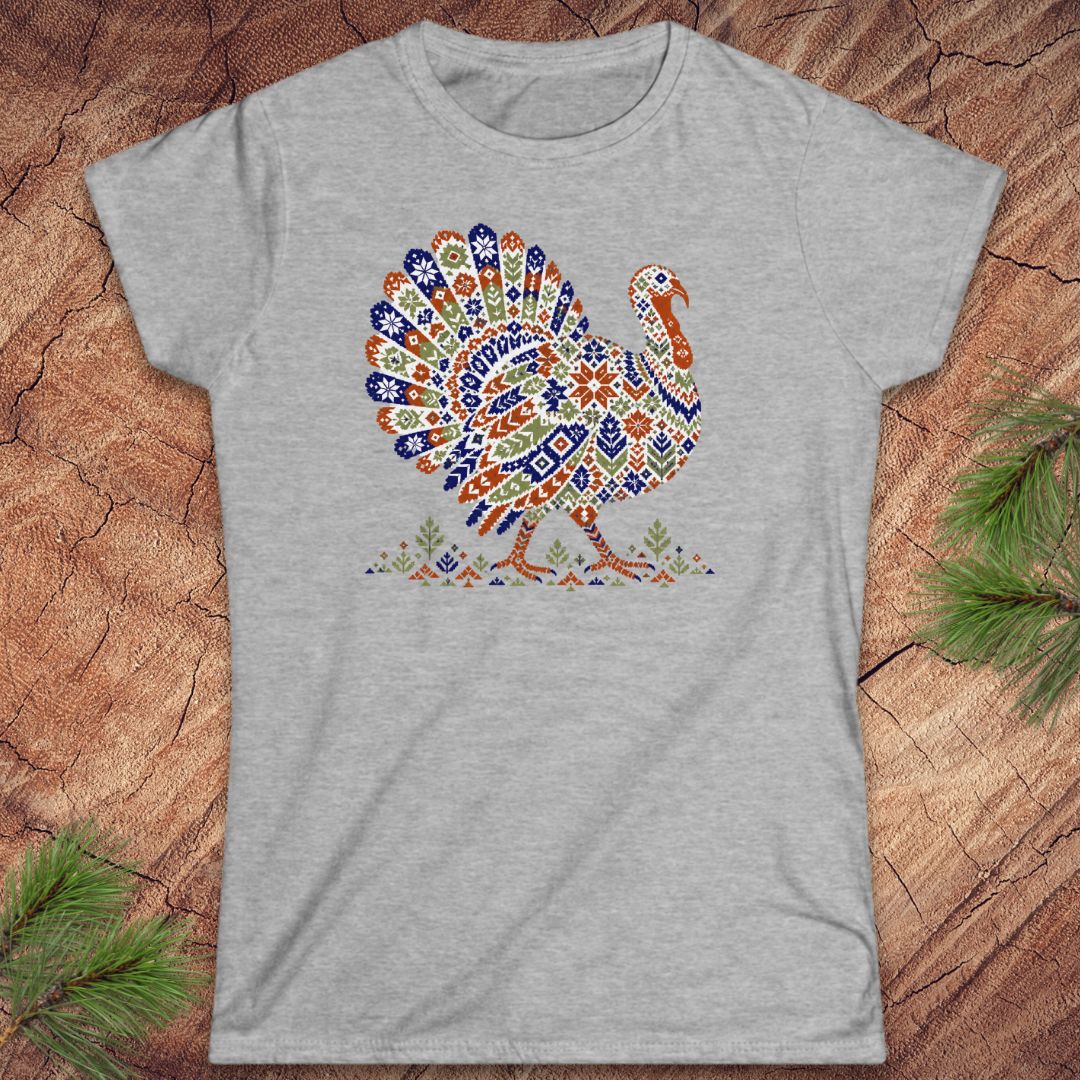 Sport grey women's t-shirt with a wild turkey patterned like a colorful knit sweater with snowflakes, stars, and tiny pine trees, laid flat on a rustic wood background with pine branches.