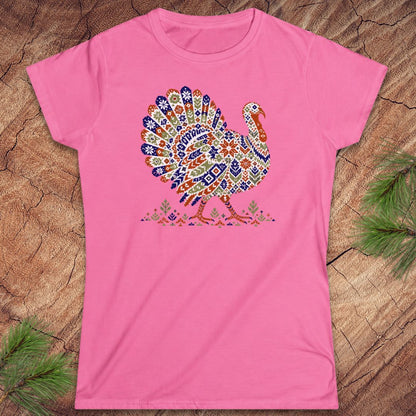 Azalea pink women's t-shirt with a wild turkey patterned like a colorful knit sweater with snowflakes, stars, and tiny pine trees, laid flat on a rustic wood background with pine branches.