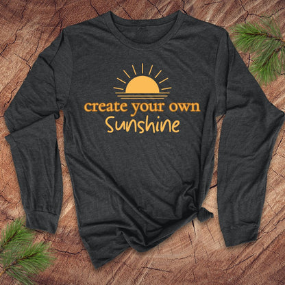 Dark grey long-sleeve tshirt with 'create your own sunshine' text and sun graphic on a wooden surface.