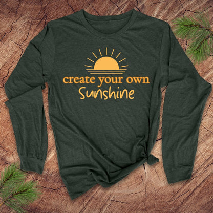 Green long-sleeve tshirt with 'create your own sunshine' text and sun graphic on a wooden surface.