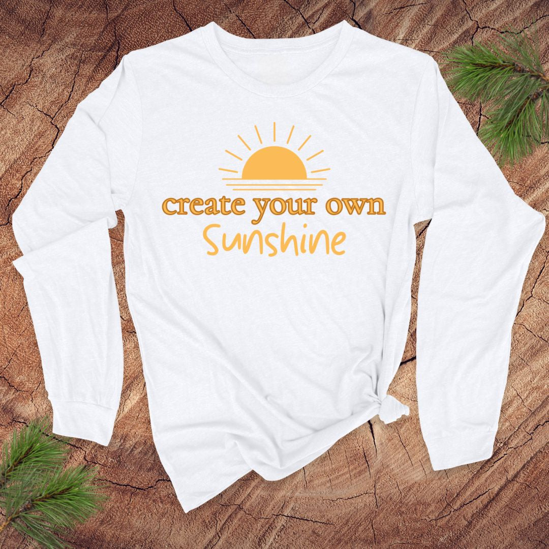 White long-sleeve tshirt with 'create your own sunshine' text and sun graphic on a wooden surface.