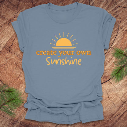 Light blue t-shirt with 'create your own Sunshine' text and sun graphic on a wooden surface.