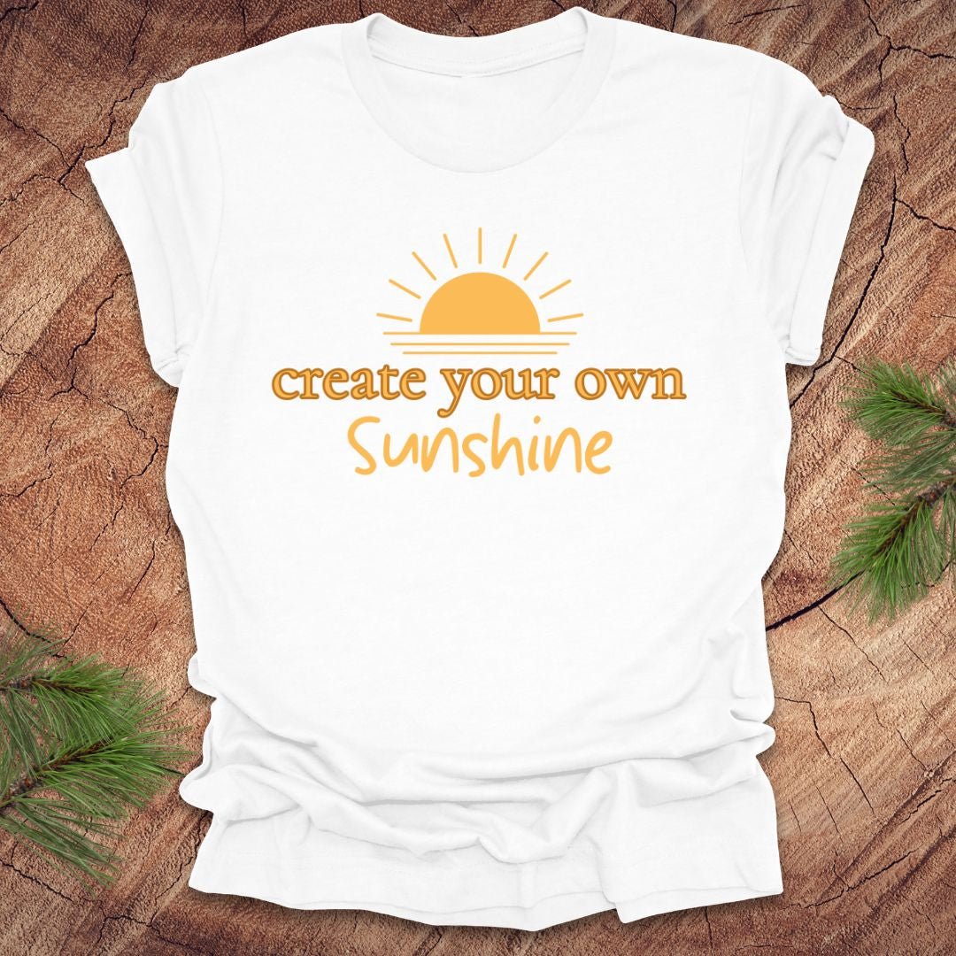 White t-shirt with 'create your own sunshine' text and sun graphic on a wooden surface.