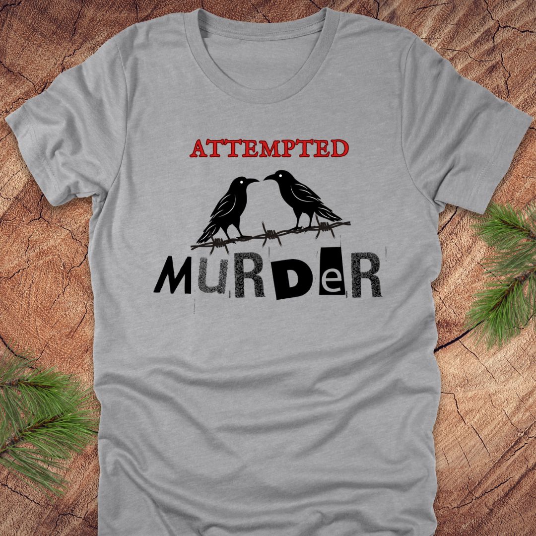 Gray t-shirt with black and red text 'Attempted Murder' and crows on a wooden surface.
