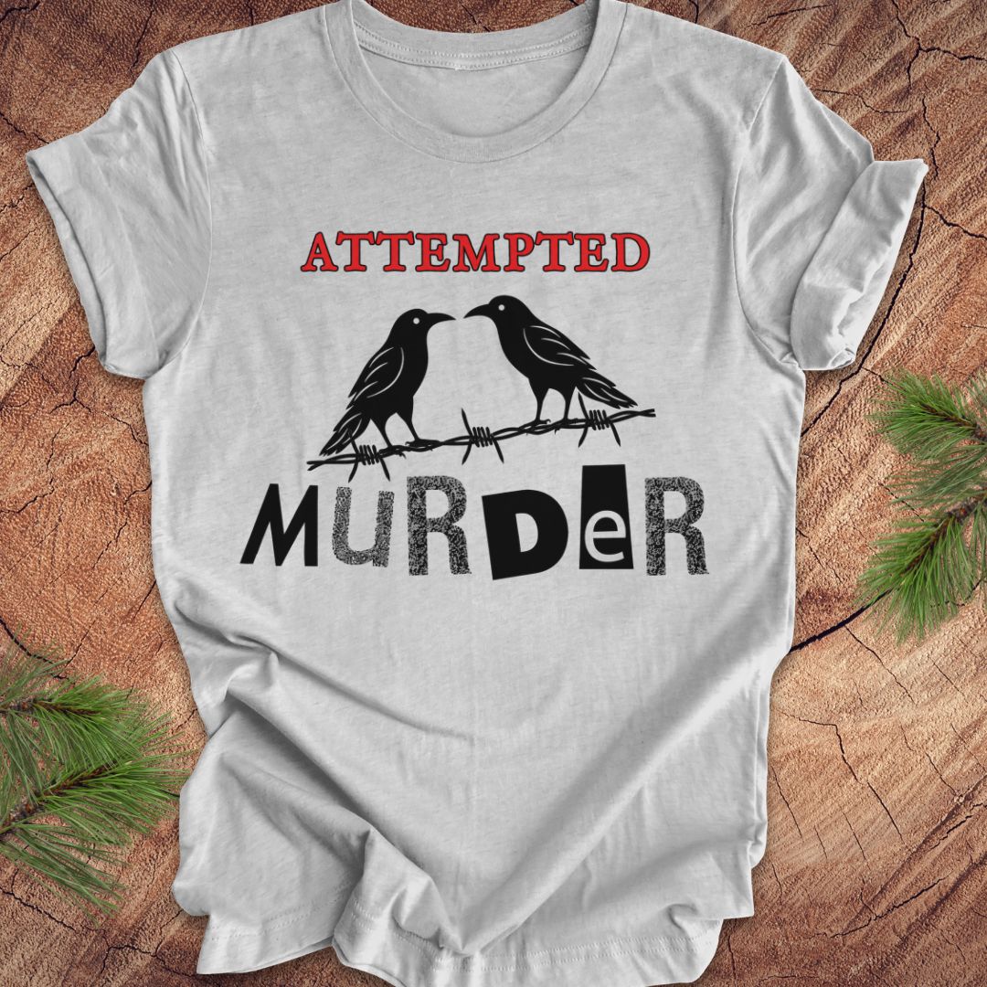 A light gray short-sleeve t-shirt with a graphic print featuring two black crows on a barbed wire with the text 'ATTEMPTED MURDER' in red and black, laid flat on a rustic wood background with pine branches.