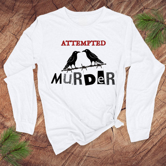 White long-sleeve shirt with black crows and text 'attempted murder' on a wooden surface