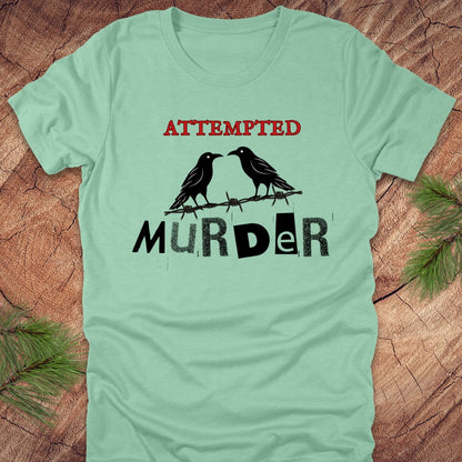 Mint green t-shirt with black and red text 'Attempted Murder' and crows on a wooden surface.