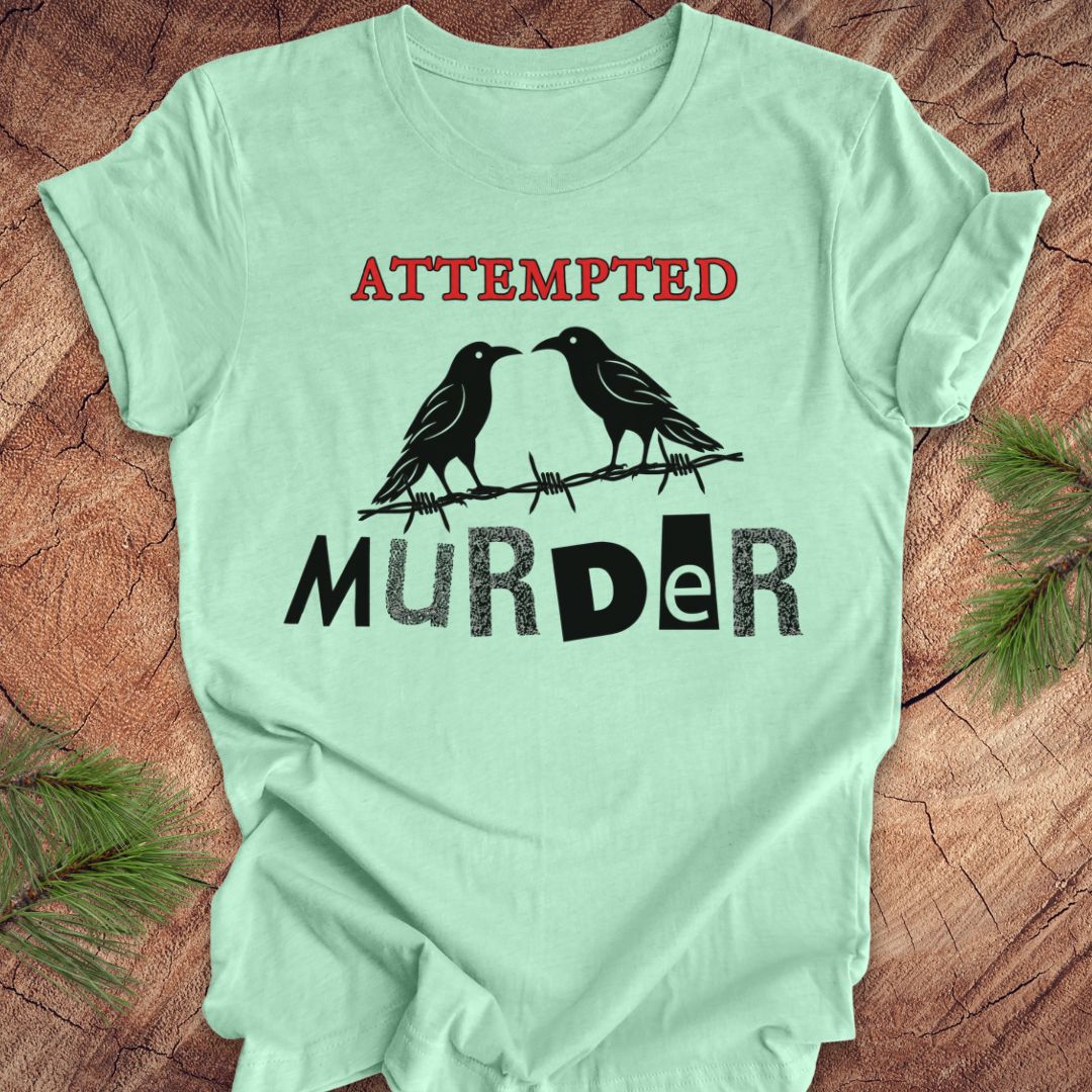 A mint green short-sleeve t-shirt with a graphic print featuring two black ravens on a barbed wire with the words 'ATTEMPTED MURDER' in red and black, laid flat on a rustic wood background with pine needles in the corners.