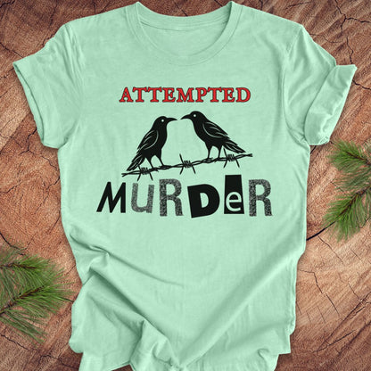 A mint green short-sleeve t-shirt with a graphic print featuring two black ravens on a barbed wire with the words 'ATTEMPTED MURDER' in red and black, laid flat on a rustic wood background with pine needles in the corners.