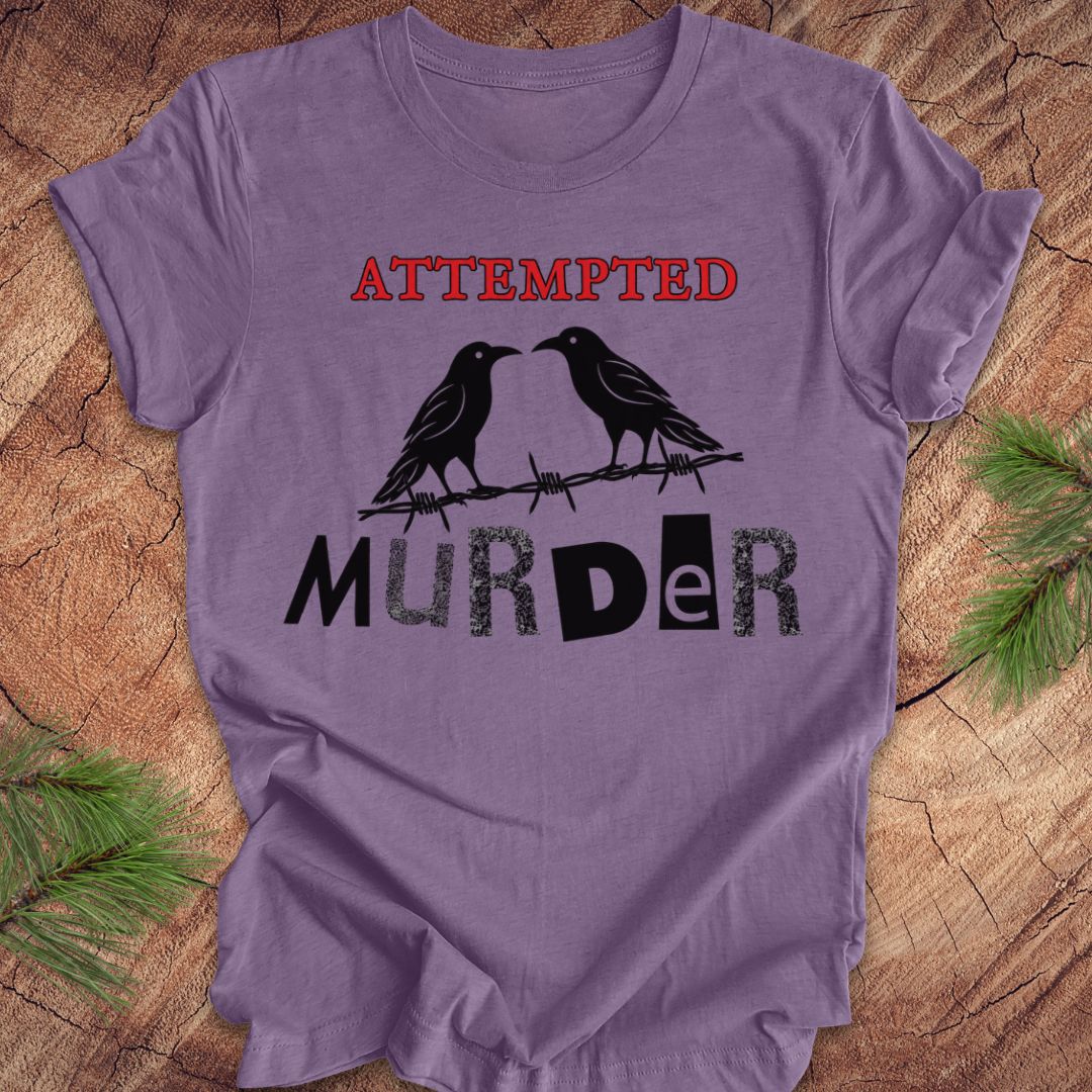 A lavender purple short-sleeve t-shirt with a graphic print reading 'ATTEMPTED MURDER' in red and black, featuring two black crows on a barbed wire. The shirt is laid flat on a rustic wood background with pine needles in the corners.