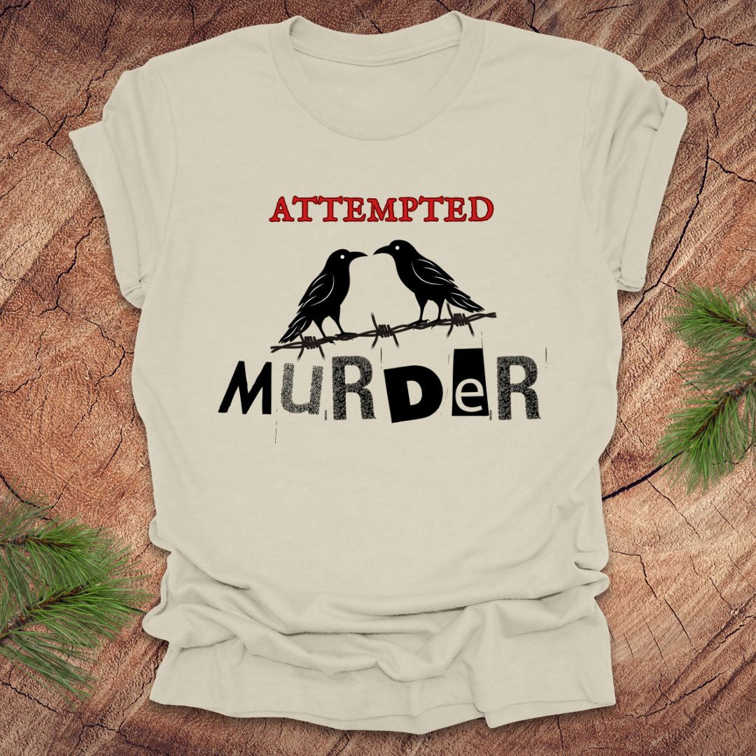 Beige t-shirt with black and red text 'Attempted Murder' and crows on a wooden surface.
