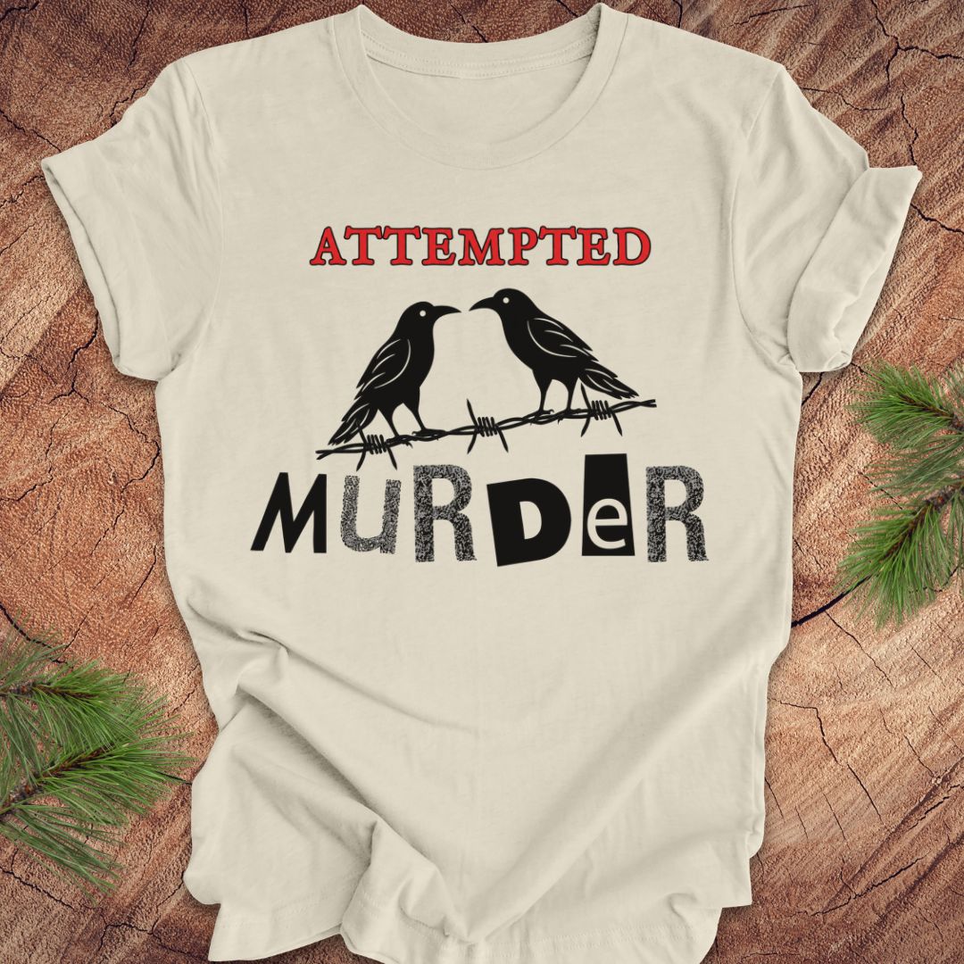 An off-white short-sleeve t-shirt with a graphic print featuring two black ravens on barbed wire and the words 'ATTEMPTED MURDER' in red and black, laid flat on a rustic wood background with pine branches.
