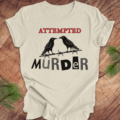 An off-white short-sleeve t-shirt with a graphic print featuring two black ravens on barbed wire and the words 'ATTEMPTED MURDER' in red and black, laid flat on a rustic wood background with pine branches.