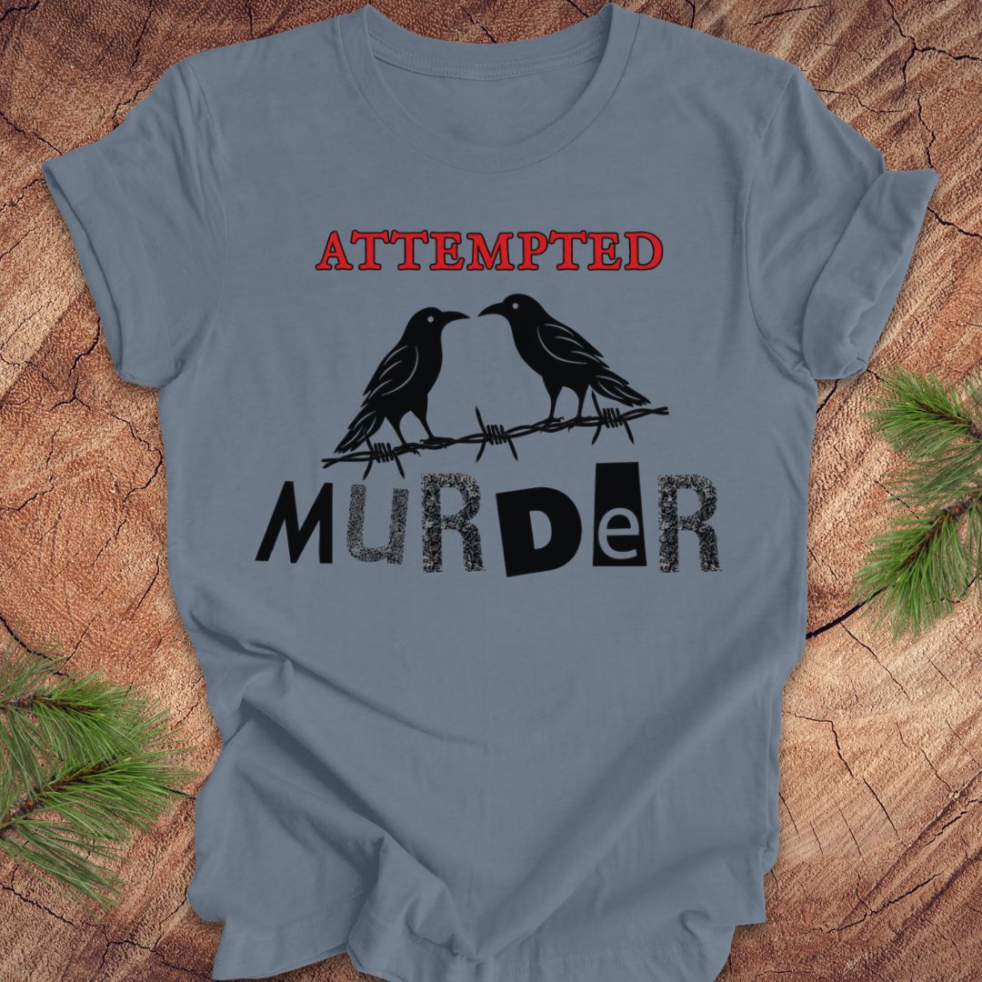 A blue crewneck t-shirt laid flat on a rustic wood background with pine branches in the corners. The shirt features a graphic print of two black ravens perched on a barbed wire with the words 'ATTEMPTED' in red above and 'MURDER' in black below.