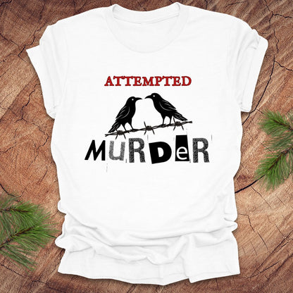 White t-shirt with black and red text 'Attempted Murder' and crows on a wooden surface.