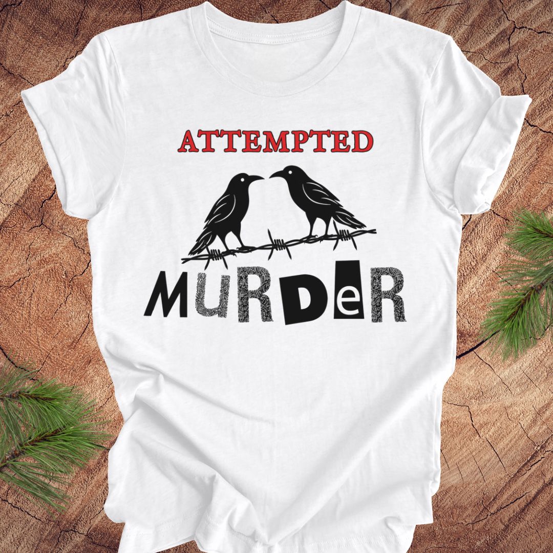 A white short-sleeve T-shirt laid flat on a rustic wood surface, printed with the words 'ATTEMPTED MURDER' in red and black, with two black birds perched on barbed wire.