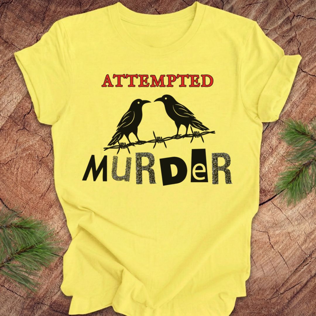 A yellow short-sleeve t-shirt laid flat on a rustic wood surface with pine branches at the corners. The shirt features a graphic print with the word 'ATTEMPTED' in red above two black crows on barbed wire, and 'MURDER' in black below.