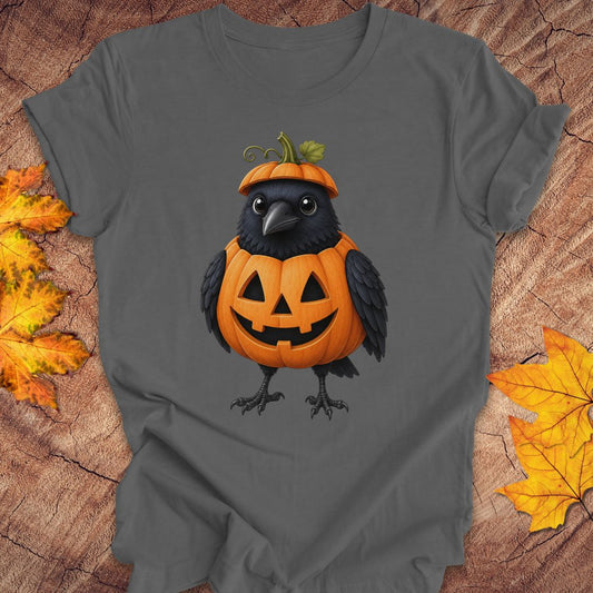 A dark gray short-sleeve t-shirt featuring a graphic of a black crow wearing an orange jack-o'-lantern pumpkin hat, styled on a rustic wood background with autumn leaves.