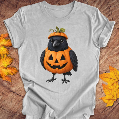 A light gray short-sleeve t-shirt with a graphic of a cute black crow wearing an orange jack-o'-lantern pumpkin hat, displayed on a rustic wood background with autumn leaves.