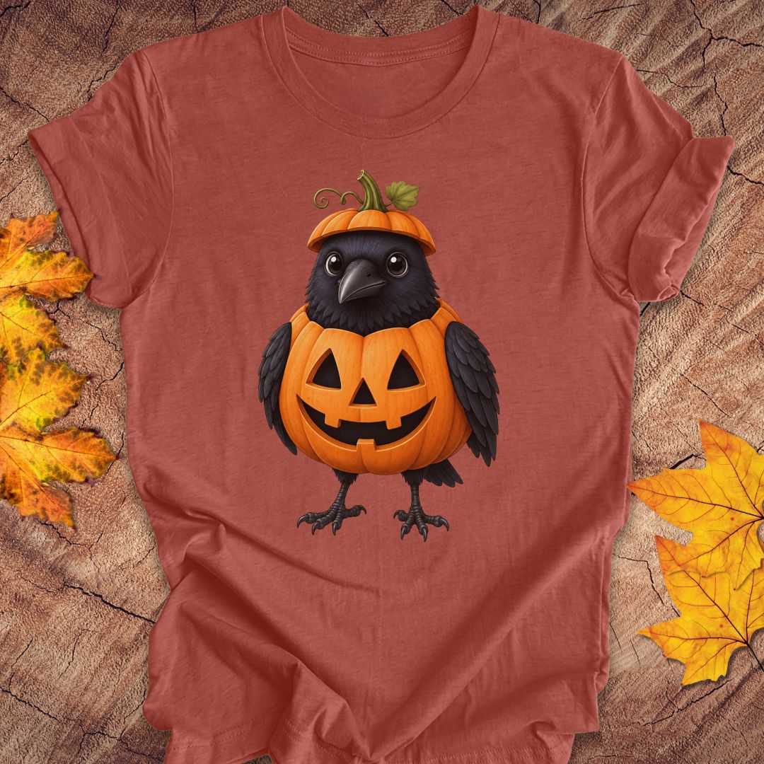 A rust-orange short-sleeve t-shirt with a graphic of a black crow wearing an orange jack-o'-lantern pumpkin hat, laid flat on a rustic wood background with yellow autumn leaves.