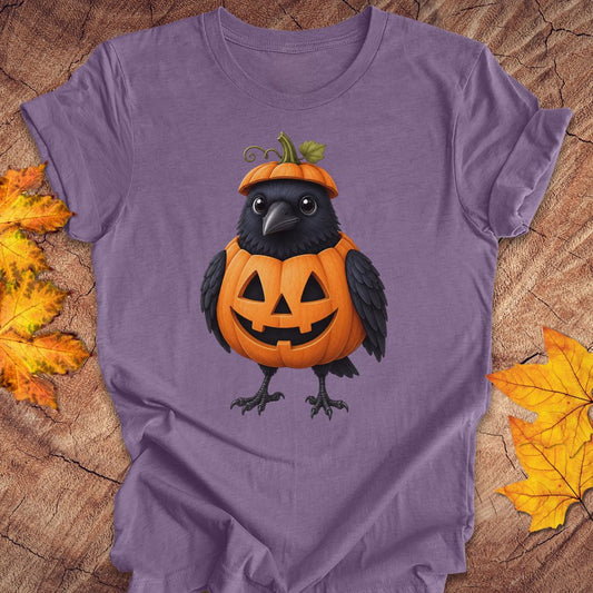 A purple short-sleeve crewneck t-shirt featuring a graphic of a cute black crow wearing an orange jack-o'-lantern pumpkin hat, laid flat on a rustic wood background with autumn leaves.