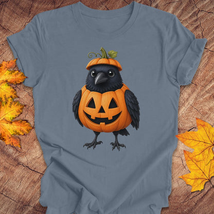 A slate blue short-sleeve t-shirt featuring a graphic of a black crow perched on a carved orange jack-o'-lantern pumpkin. The t-shirt is laid flat on a rustic wood background, framed by decorative yellow and orange autumn leaves in the corners.