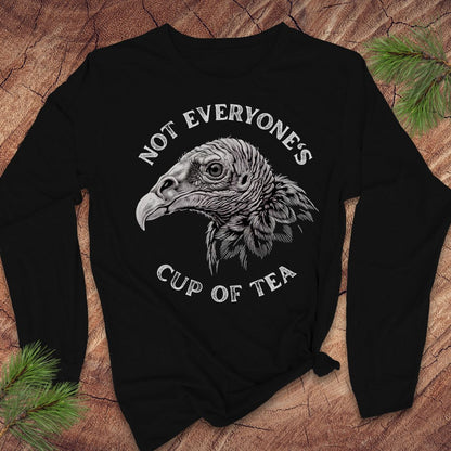 A black long-sleeve t-shirt with a white graphic of a vulture head and the text 'NOT EVERYONE'S CUP OF TEA', laid flat on a rustic wood background with pine branches.