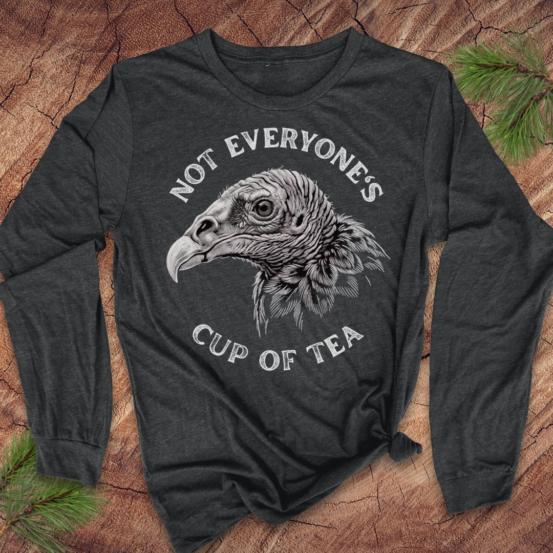 A dark charcoal gray long-sleeve t-shirt with a graphic of a vulture head and the text 'NOT EVERYONE'S CUP OF TEA' in white. The shirt is laid flat on a rustic wood background with pine needles in the corners.
