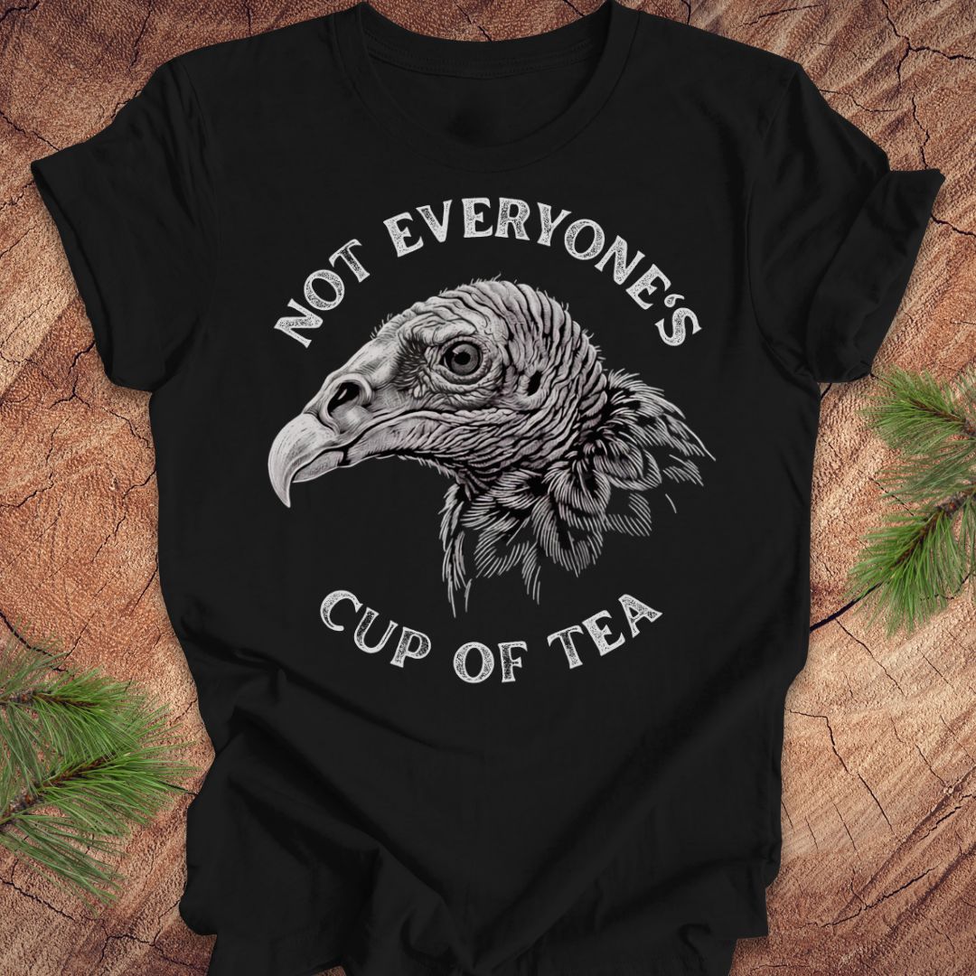 A black short-sleeve graphic T-shirt laid flat on a rustic wood surface with pine branches at the corners. The shirt features a large white and gray vulture head illustration in the center, with the words 'NOT EVERYONE'S CUP OF TEA' curved above and below the graphic.