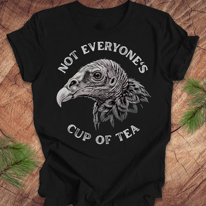 A black short-sleeve graphic T-shirt laid flat on a rustic wood surface with pine branches at the corners. The shirt features a large white and gray vulture head illustration in the center, with the words 'NOT EVERYONE'S CUP OF TEA' curved above and below the graphic.
