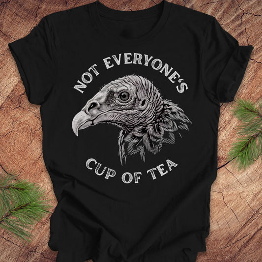 A black short-sleeve graphic T-shirt laid flat on a rustic wood surface with pine branches at the corners. The shirt features a large white and gray vulture head illustration in the center, with the words 'NOT EVERYONE'S CUP OF TEA' curved above and below the graphic.