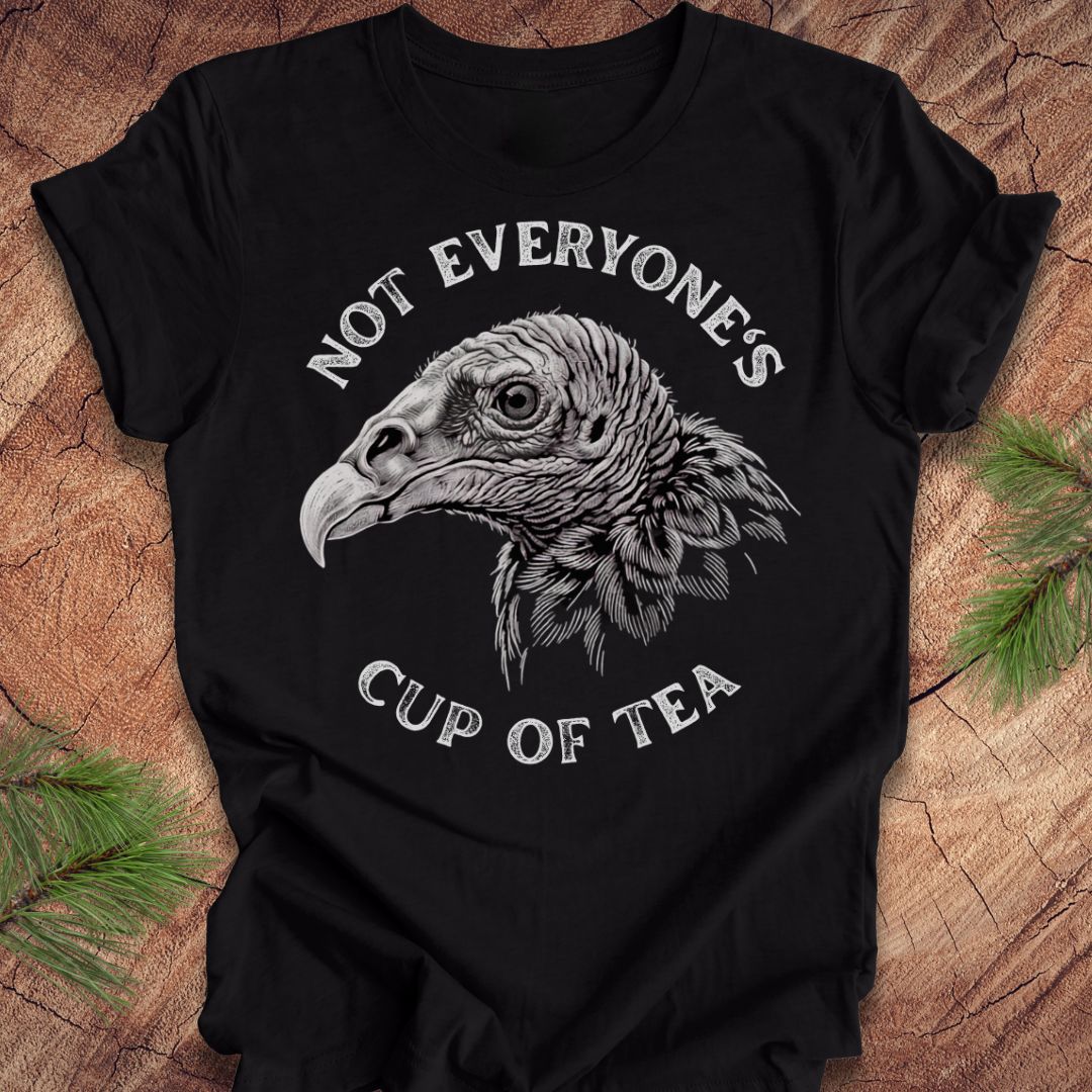 A black short-sleeve t-shirt with a detailed white graphic of a vulture's head and the text 'NOT EVERYONE'S CUP OF TEA' printed on the front, displayed flat on a rustic wood background with pine branches in the corners.