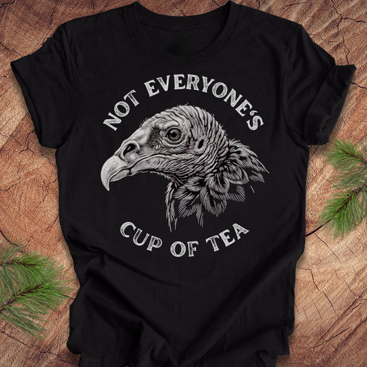 A black short-sleeve t-shirt with a detailed white graphic of a vulture's head and the text 'NOT EVERYONE'S CUP OF TEA' printed on the front, displayed flat on a rustic wood background with pine branches in the corners.