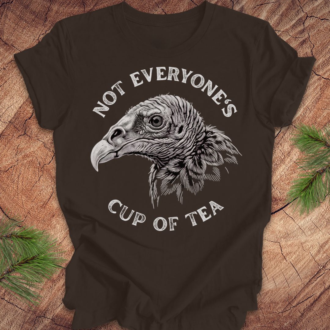 A dark brown short-sleeve t-shirt with a white graphic of a vulture head and the text 'NOT EVERYONE'S CUP OF TEA', displayed flat on a rustic wood background with pine branches.