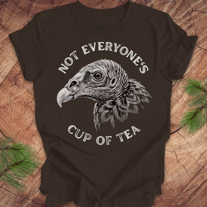 A dark brown short-sleeve t-shirt with a white graphic of a vulture head and the text 'NOT EVERYONE'S CUP OF TEA', displayed flat on a rustic wood background with pine branches.