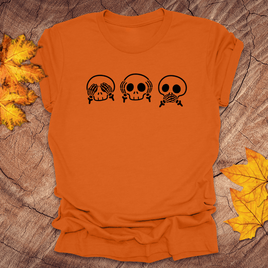 Orange tshirt with 3 skulls simulating see no evil, hear no evil, speak no evil, on a wood background with fall leaves.