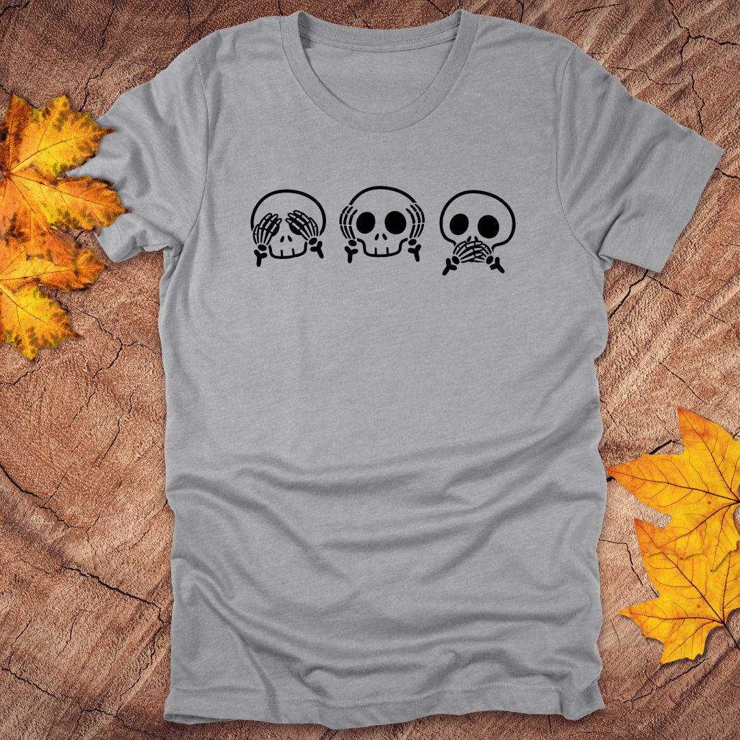 Gray tshirt with 3 skulls simulating see no evil, hear no evil, speak no evil, on a wood background with fall leaves.